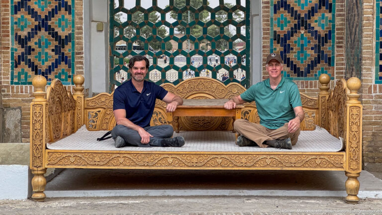 Closer portrait of two men seated on a carved wooden platform before green latticework and blue tiles.