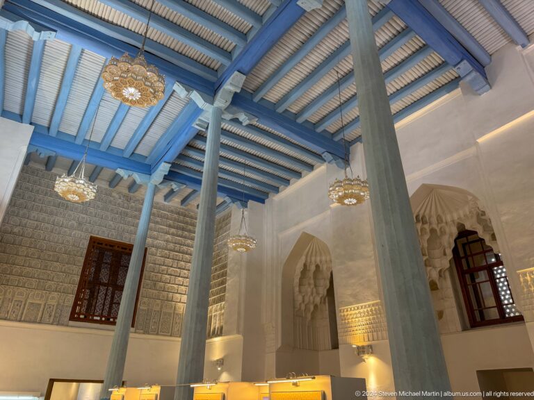 Columned interior with a blue-painted timber ceiling, chandeliers, carved niches, and screened windows.
