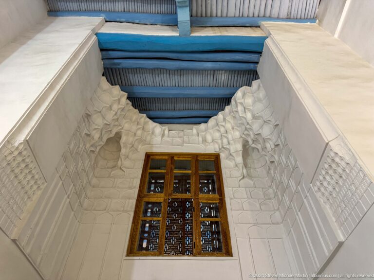 Narrow white plaster interior with blue ceiling beams and a small wooden lattice window.