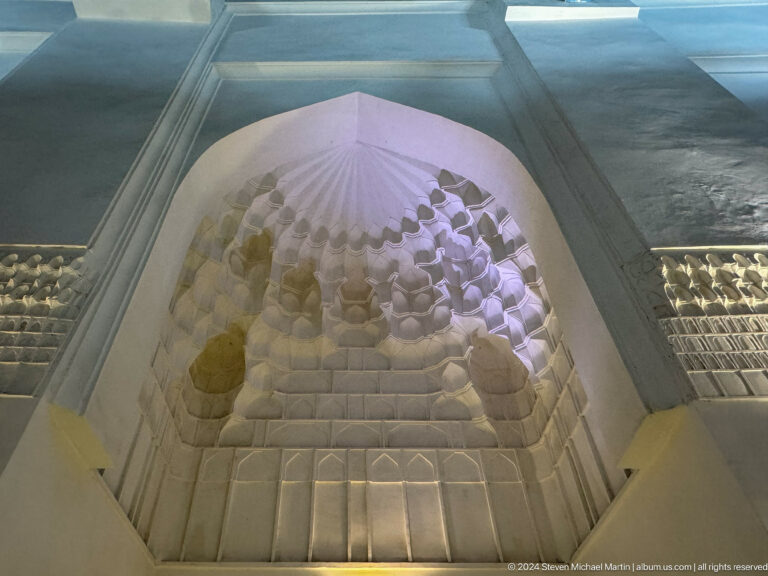 Upward view into a tall white plaster muqarnas niche softly lit violet.