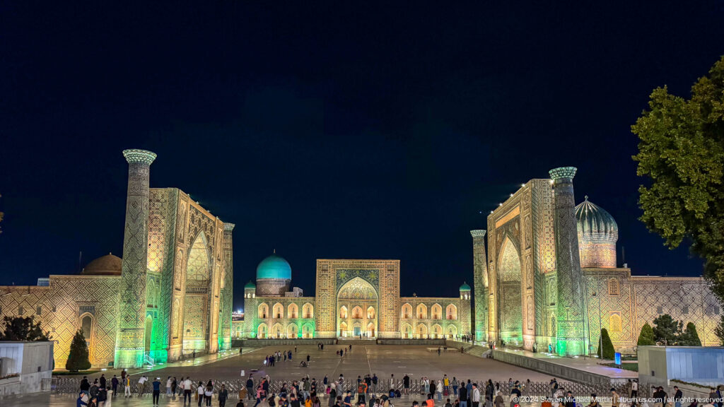 Elevated nighttime panorama of all three Registan madrasas illuminated in white and green.