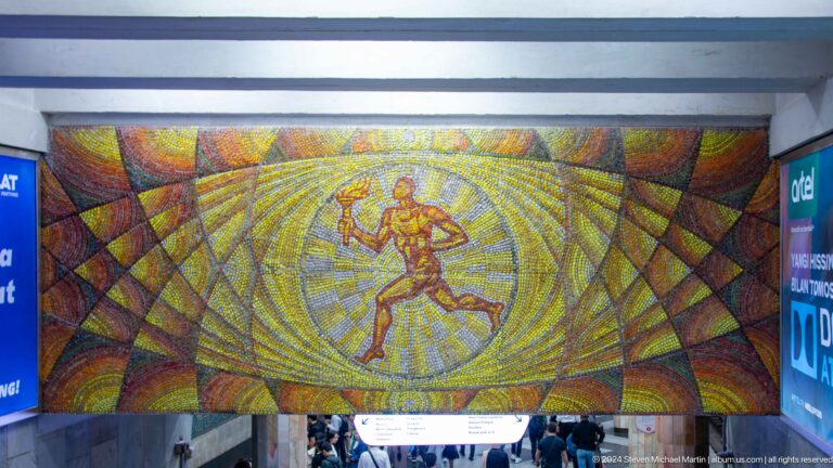 Large mosaic of a running torchbearer above a crowded metro concourse in Tashkent.
