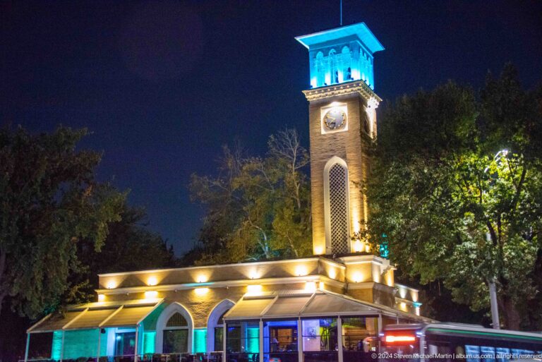 Illuminated clock tower rising above a low pavilion with arched openings at night.