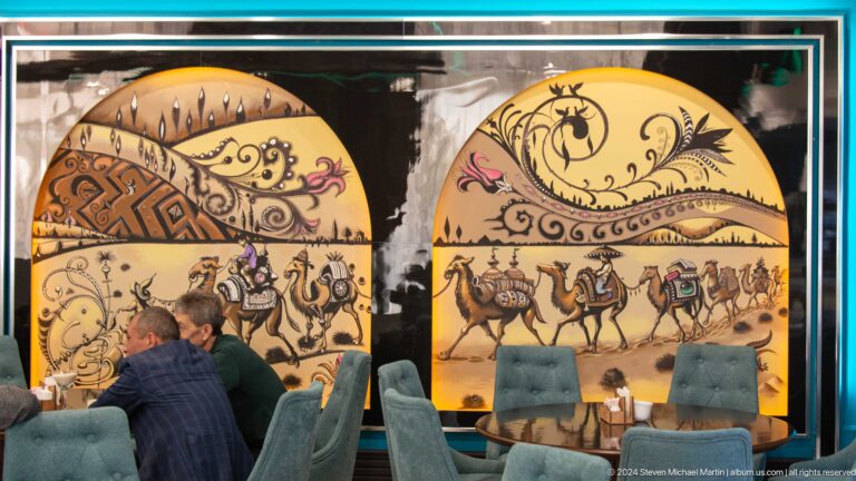 Arched hotel mural showing musicians, mounted travelers, camels, and ornamental landscape motifs.