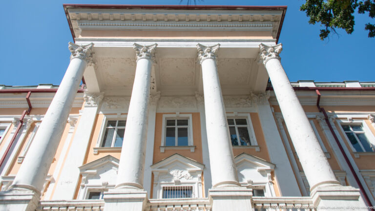 Low-angle view of a peach facade with four tall white columns and ornate capitals.