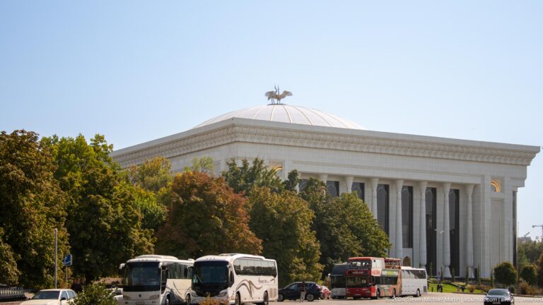 Large colonnaded building with a ribbed dome and bird sculpture behind tour buses and trees.