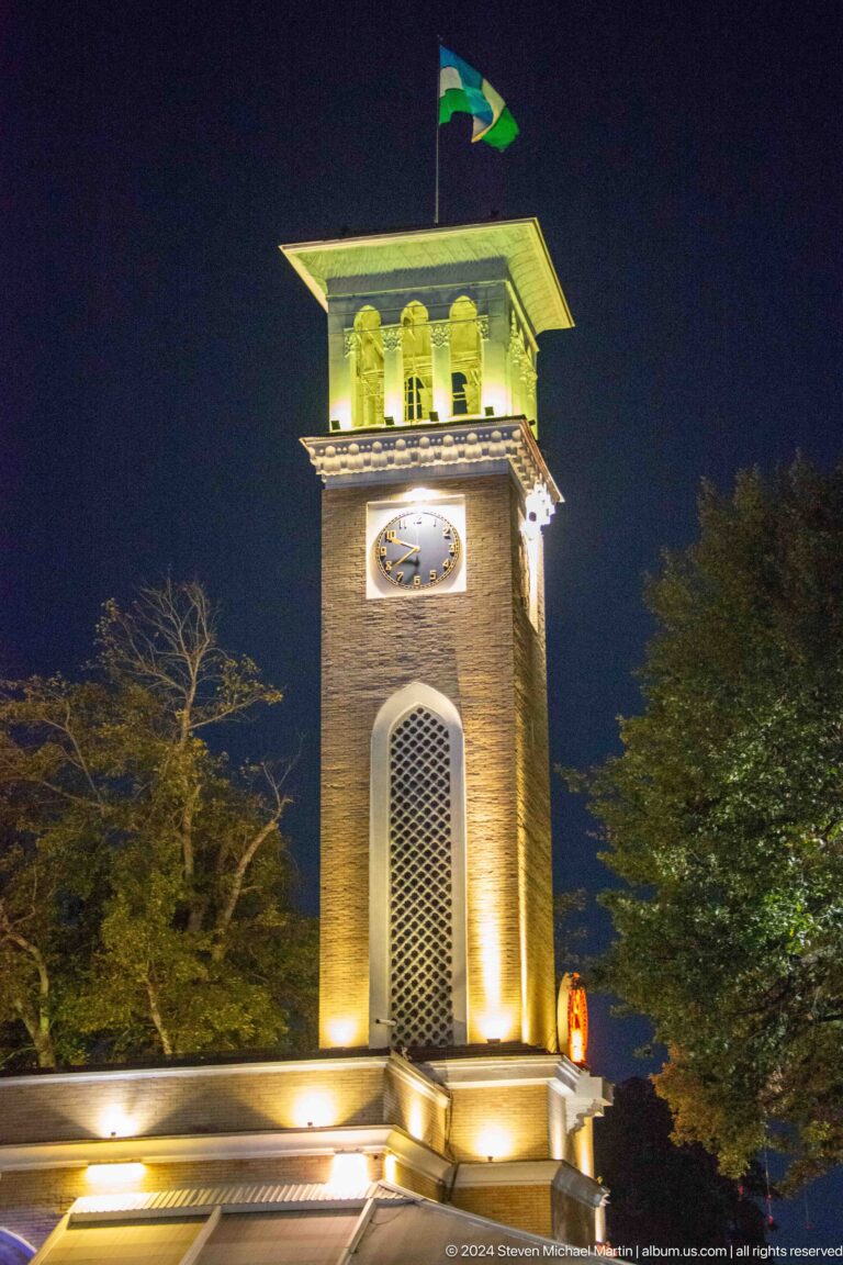Vertical night view of a masonry clock tower with illuminated face and pointed-arch opening.