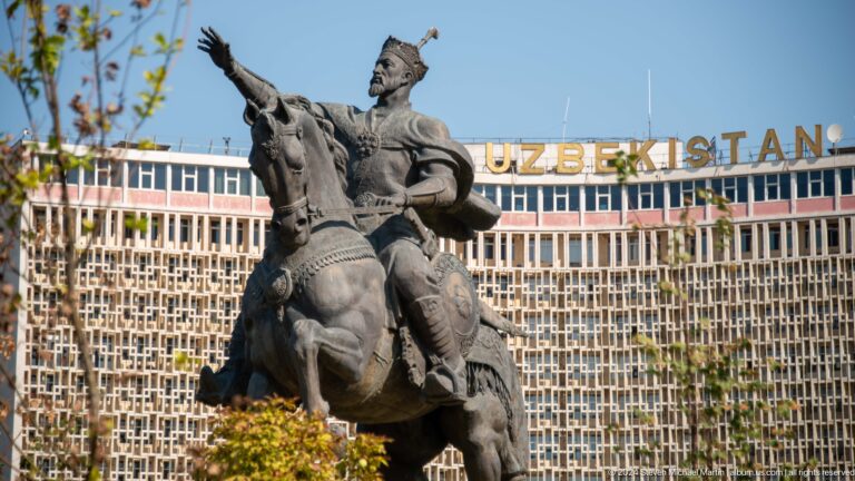 Full Amir Timur equestrian statue with Hotel Uzbekistan's geometric facade behind.