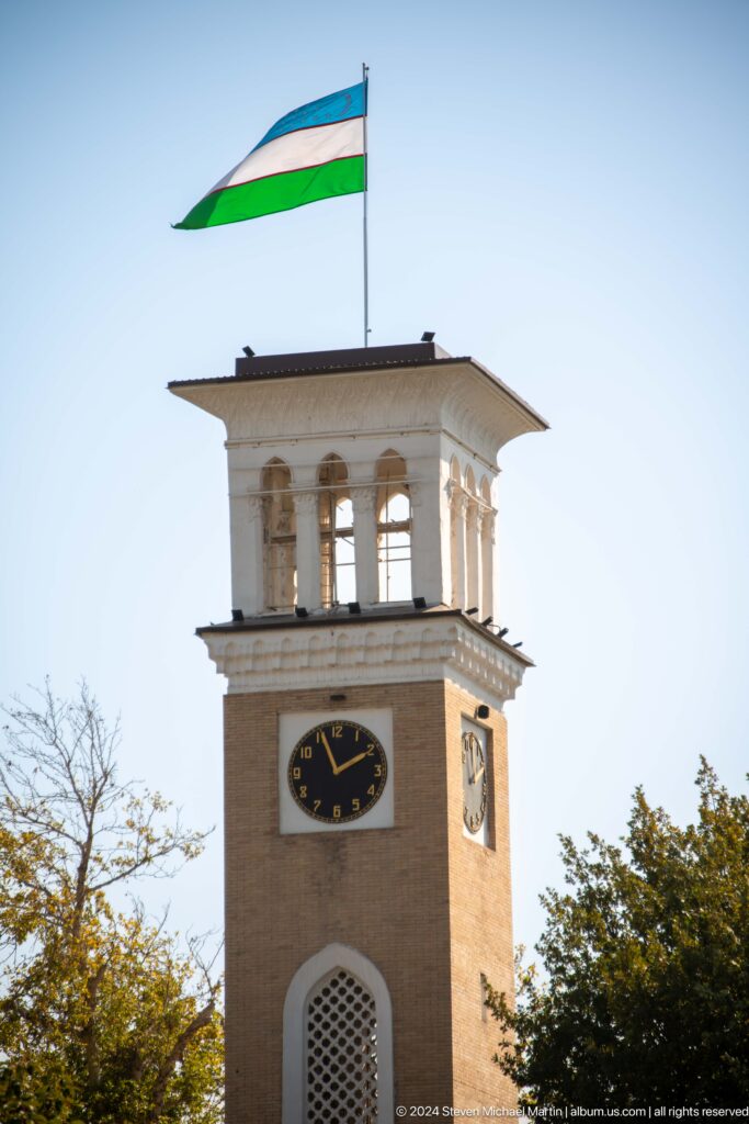 Tan brick clock tower with a black clock face and open white pavilion at the top.