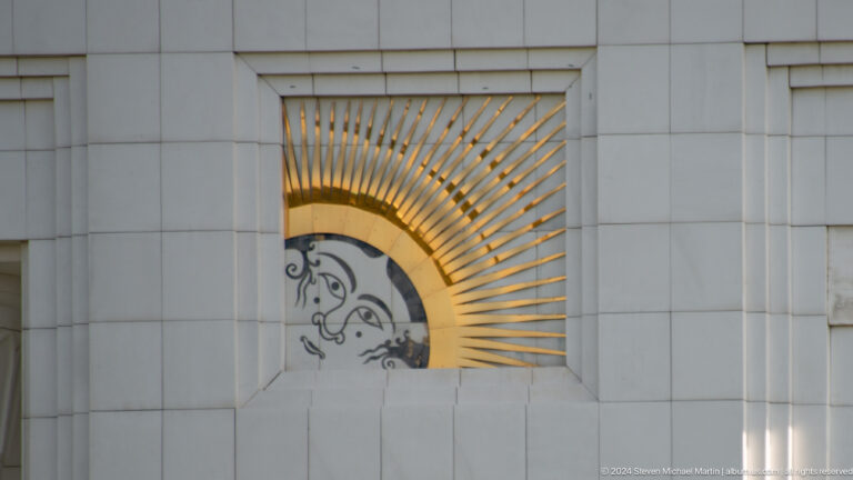 Arched wall panel with radiating gold rods surrounding a stylized black figure.