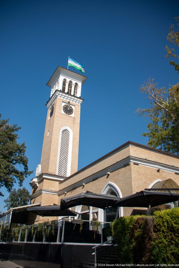 Full tan brick clock tower with white lattice panel beside a low public building.