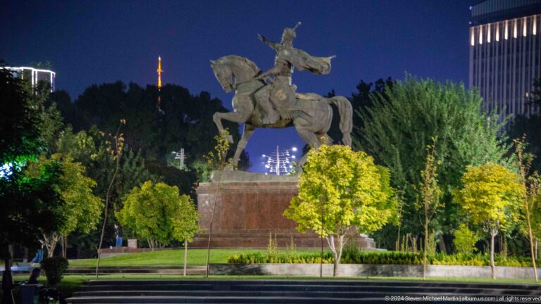 Side view of the Amir Timur equestrian monument in a landscaped square at night.