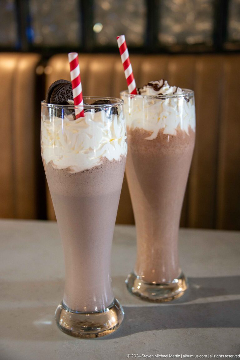 Two tall chocolate milkshakes topped with whipped cream, one with a red cherry.
