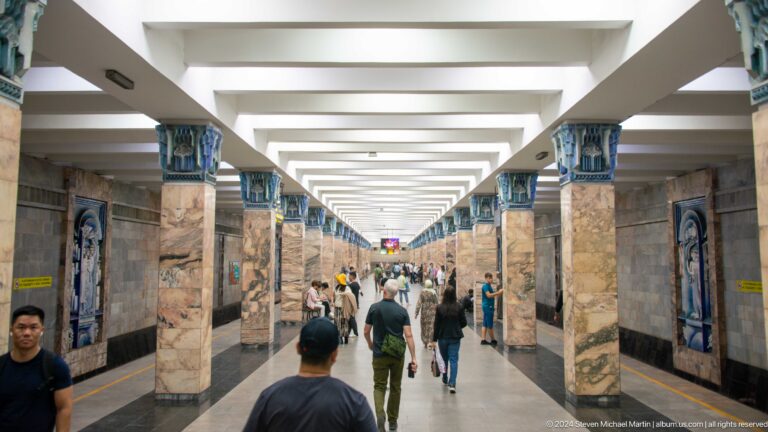 Long Toshkent Metro station platform with marble columns, blue capitals, and many passengers.