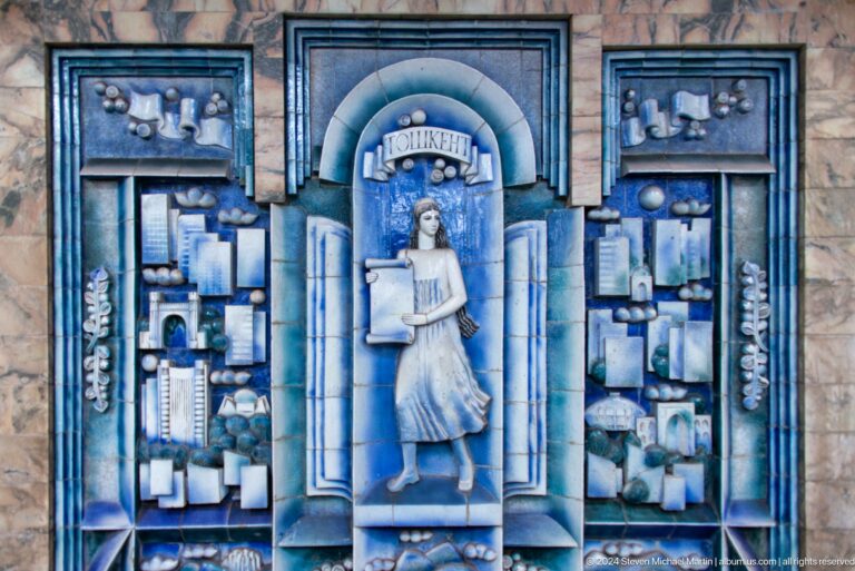 Blue ceramic relief of a woman holding scrolls amid books, domes, and city buildings.