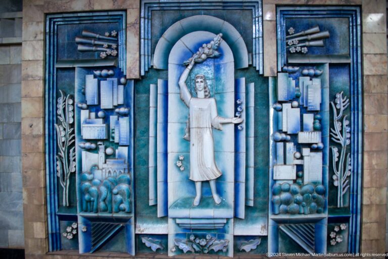 Blue ceramic relief of a woman raising a flowering branch above stylized city motifs.