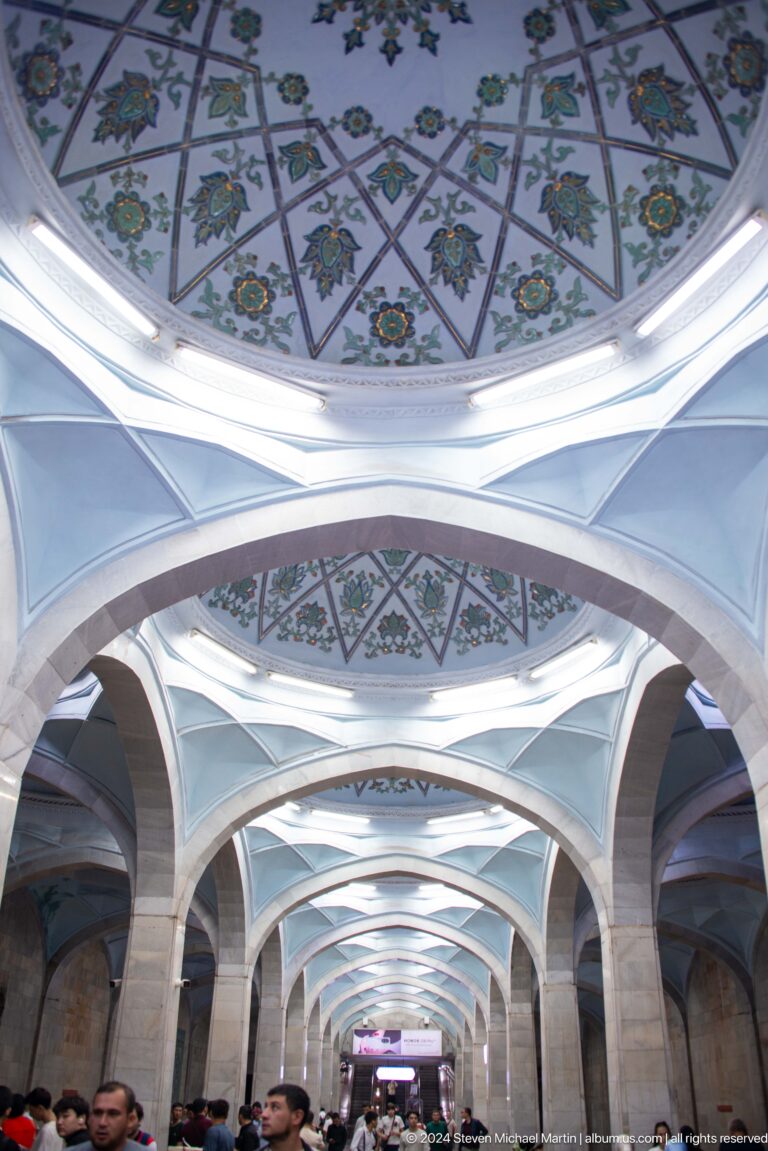 Upward view of pointed metro arches, faceted white vaults, lights, and floral painted bands.
