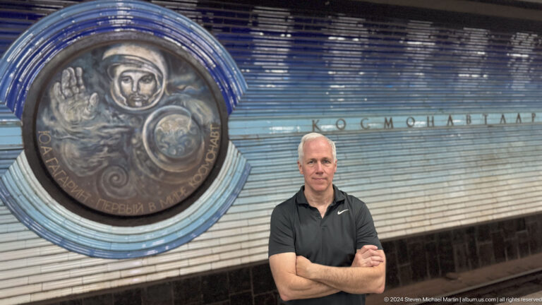 Adult male visitor with folded arms standing before a large cosmonaut relief at Kosmonavtlar station.