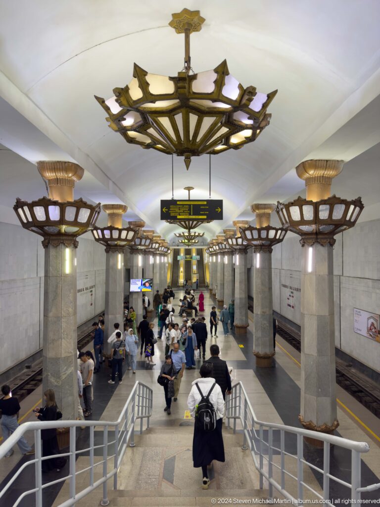 Elevated view of a crowded metro platform lined with columns and basket-shaped light fixtures.