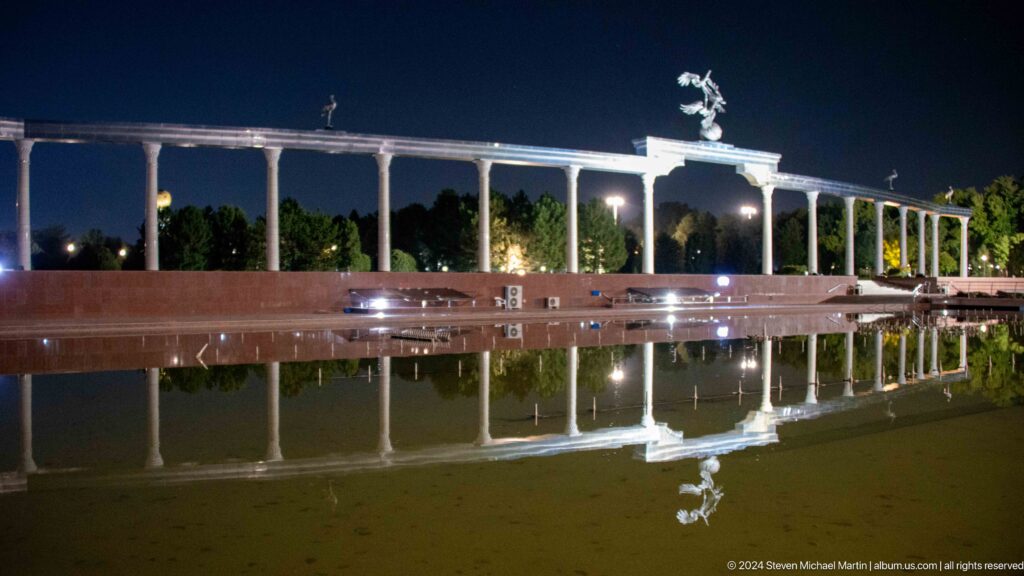 Long white memorial colonnade and bird sculpture reflected in a still pool at night.