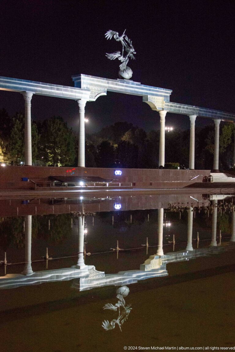 White memorial columns and a low stone wall reflected in a dark pool at night.