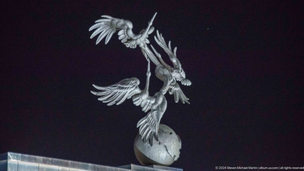 Silver sculpture of three flying storks rising from a cracked globe against the night sky.
