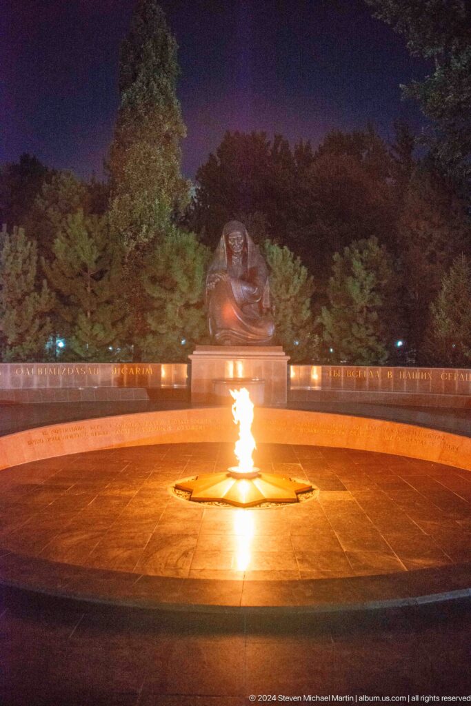 Wide night view of an eternal flame, seated grieving woman, and curved memorial wall.