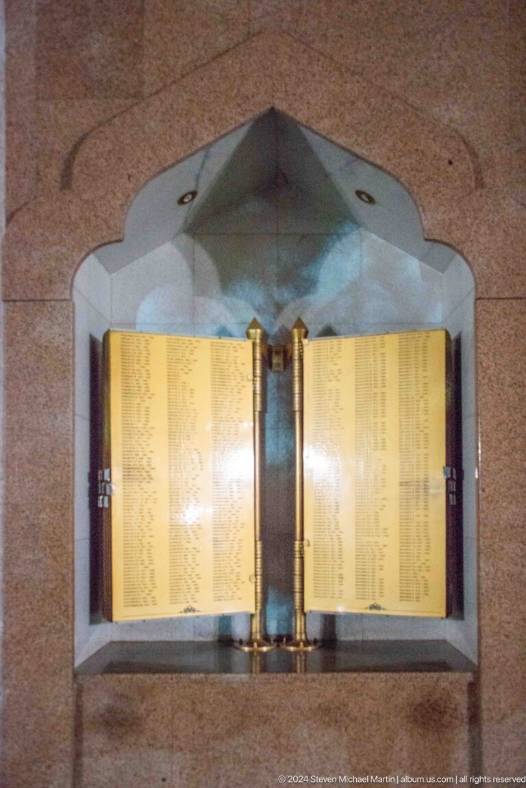 Golden open memorial book filled with columns of names inside a pointed-arch stone niche.