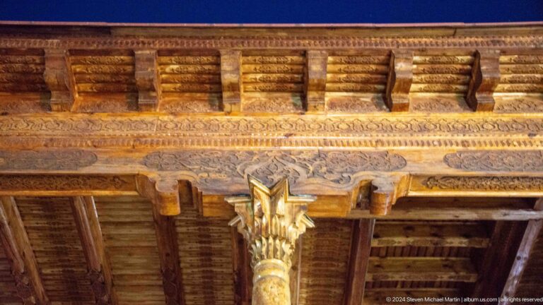 Upward view of carved timber beams and a gold-accented column capital in a memorial colonnade.