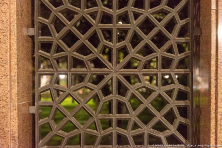 Square wooden lattice screen with interlocking diagonal strips and greenery visible beyond.