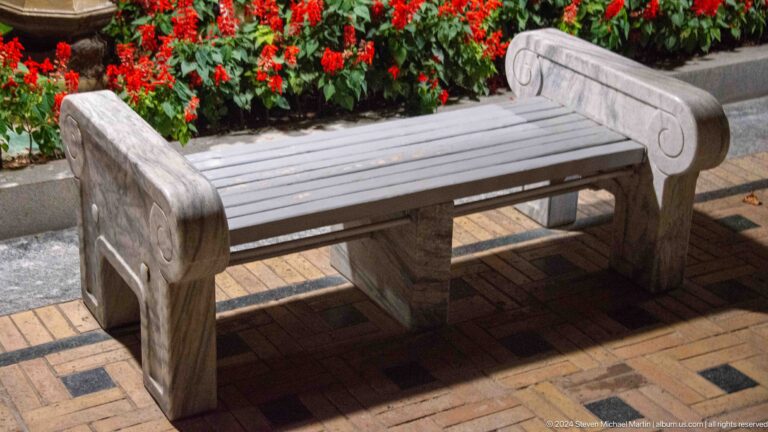 Pale carved stone bench beside a flowerbed filled with red blossoms.