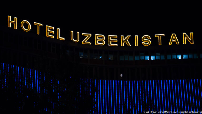 Gold HOTEL UZBEKISTAN letters glowing above blue facade lights against a black sky.