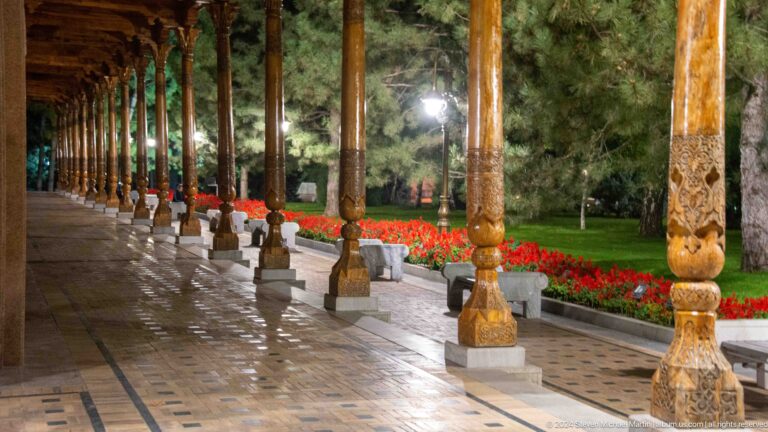 Long illuminated memorial colonnade beside red flowers and trimmed hedges at night.