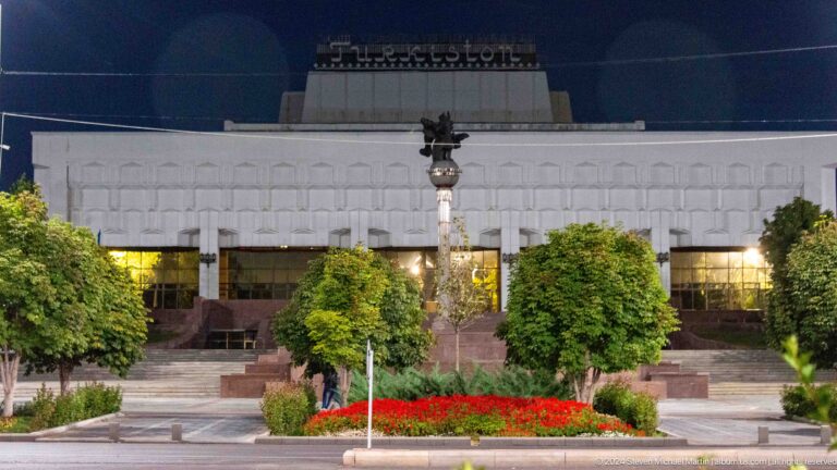 Large pale civic building behind red flowerbeds, trimmed trees, and decorative lamps at night.