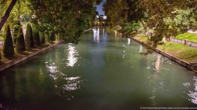 Tree-lined water channel with small fountains and reflected lights at night.