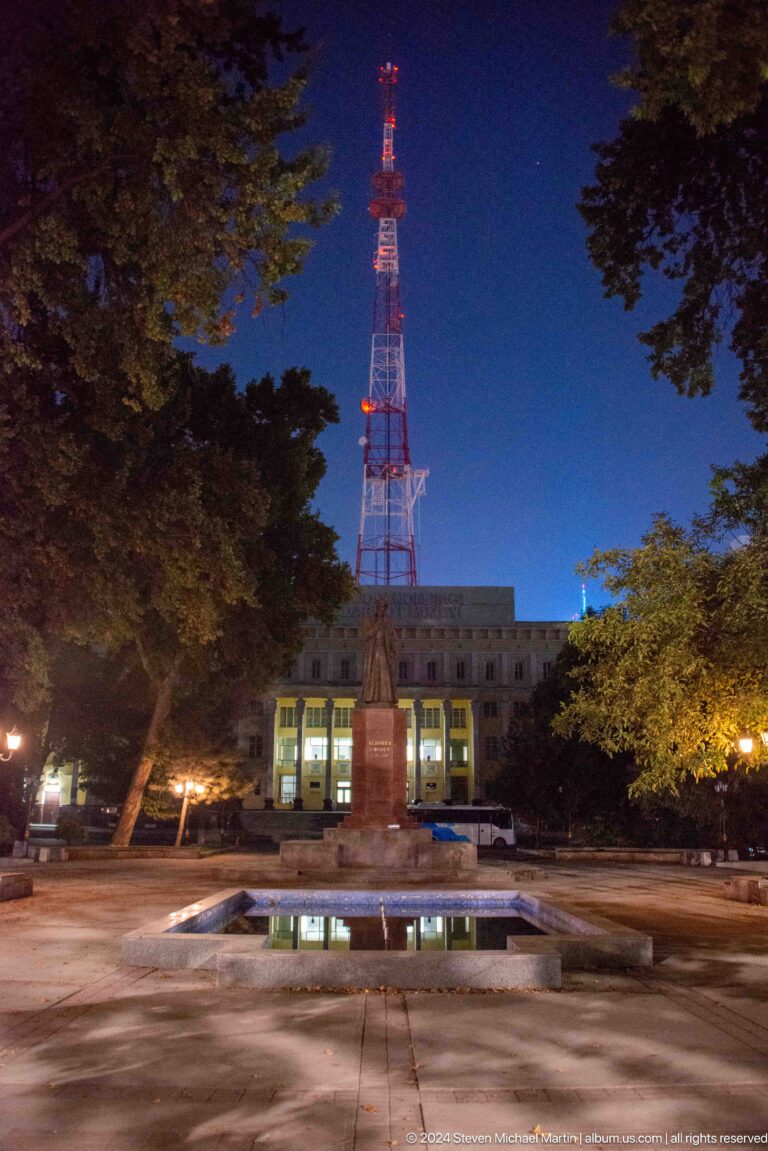 Red-and-white communications mast rising above trees and a low building at dusk.