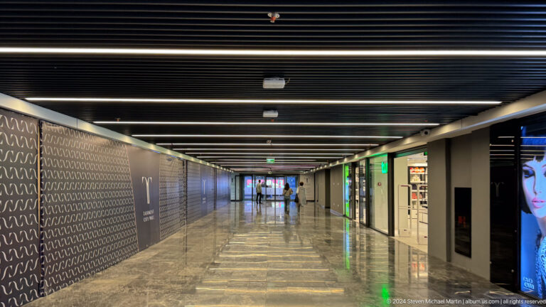 Long polished underground metro corridor with patterned walls, dark ceiling bands, and bright shopfronts.