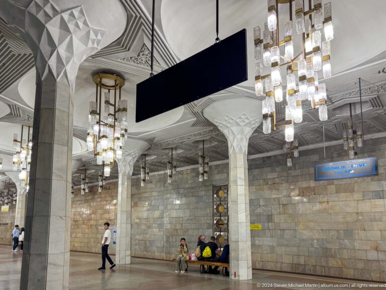 Bright metro hall with white flared columns and clusters of hanging chandeliers.