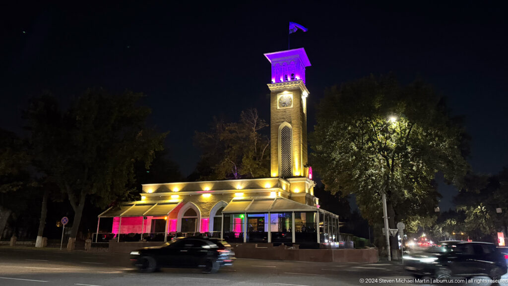 Clock tower with a purple-lit crown rising above an illuminated low pavilion at night.
