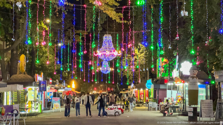 Busy pedestrian street covered with multicolored hanging lights and chandelier-shaped decorations at night.
