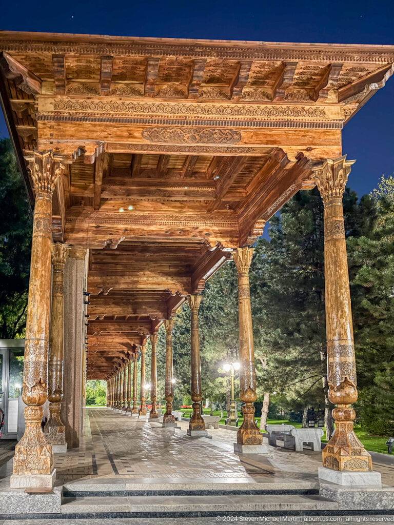 Long carved wooden memorial colonnade receding through a landscaped setting at night.