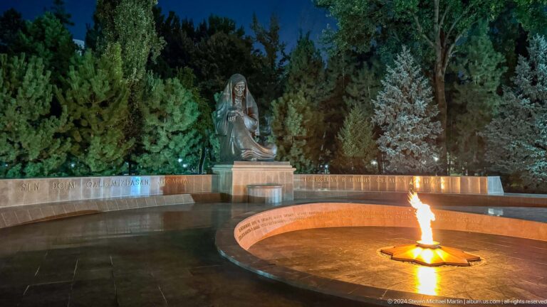 Wide night view of an eternal flame before a seated bronze grieving woman and curved memorial wall.