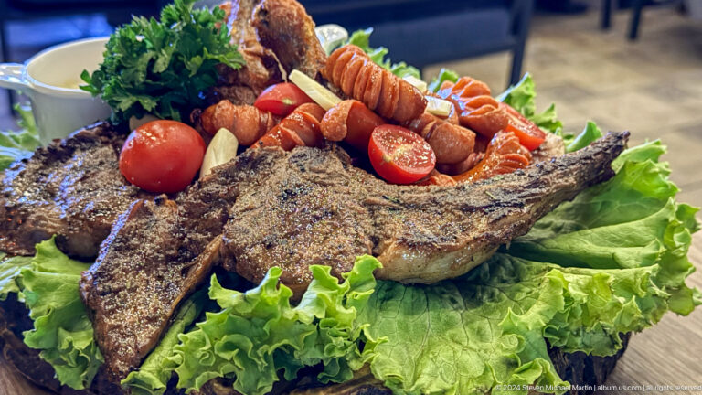 Whole grilled fish on lettuce with tomatoes, sausages, and skewered accompaniments.