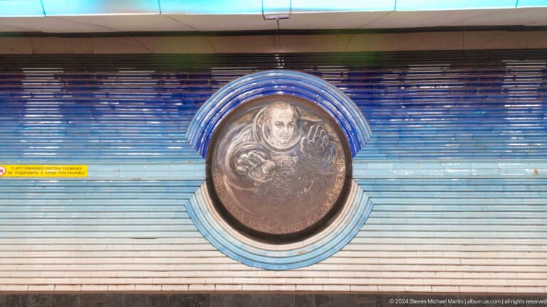 Round bronze relief of a helmeted cosmonaut raising one hand, framed by blue ceramic bands.