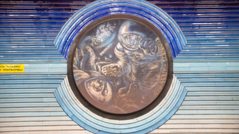 Round bronze relief of several helmeted cosmonauts reaching toward one another.