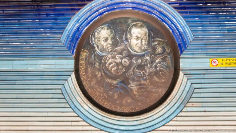 Round relief of two male cosmonauts above spacecraft instruments and symbolic motifs.