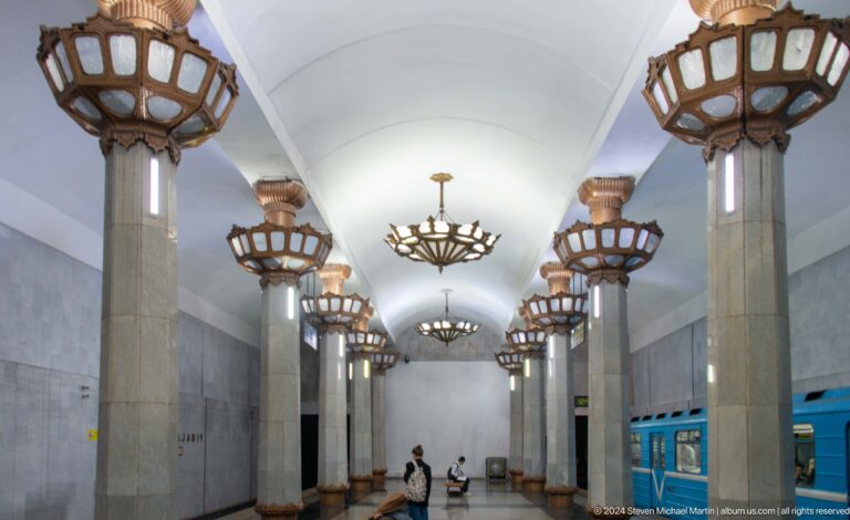 Grand metro hall with tall pale columns, basket-like light fixtures, and hanging chandeliers.