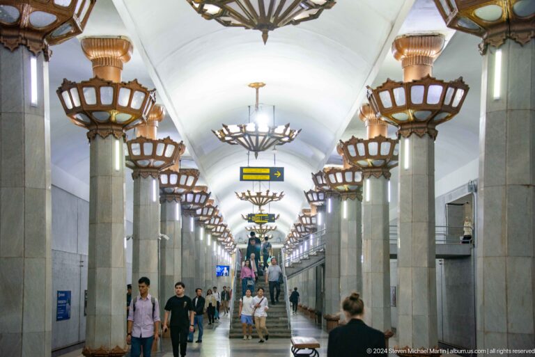 Long metro hall with pale columns, basket-shaped illuminated capitals, and a white vaulted ceiling.