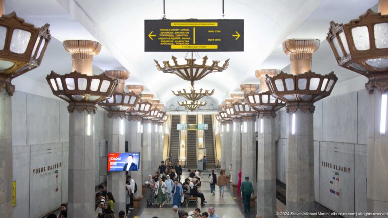 Busy Tashkent Metro concourse with passengers moving toward escalators beneath a direction sign.