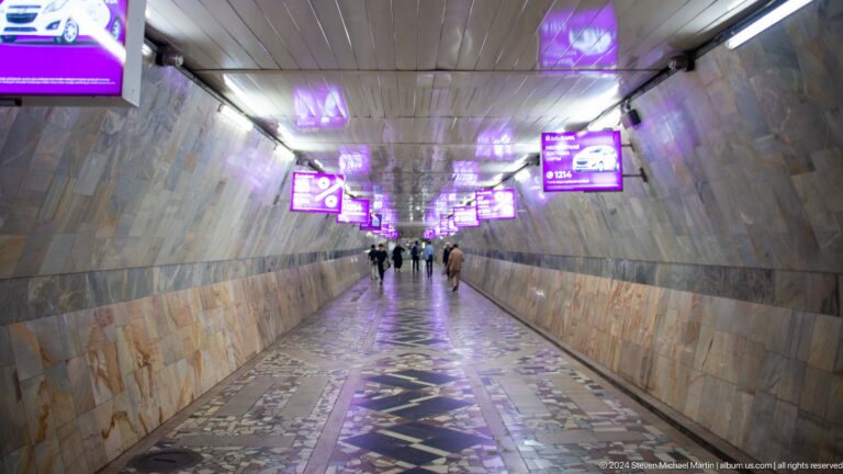 Long marble-lined metro tunnel with purple advertising screens and a patterned reflective floor.